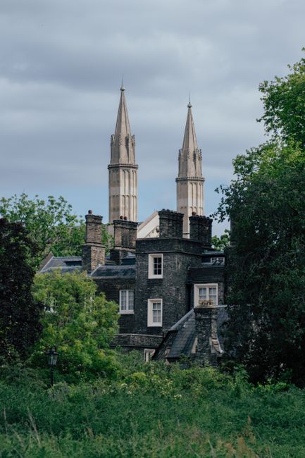 A historic stone-built building with multiple chimneys and a slate roof, partially obscured by lush green trees in the foreground. The building features small, rectangular windows and appears to be si