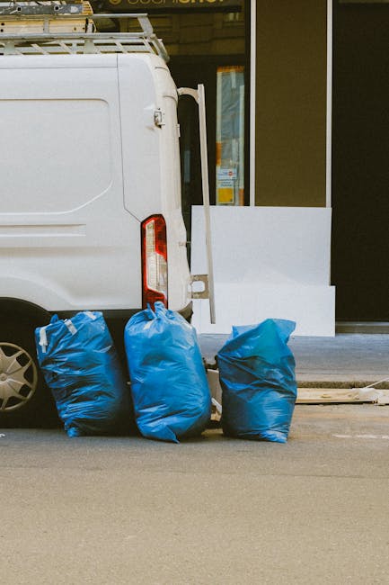 A narrow urban alleyway cluttered with a large, wheeled black rubbish bin in the foreground, which appears to be made of durable plastic with a textured surface and is partially covered in dirt. Behin