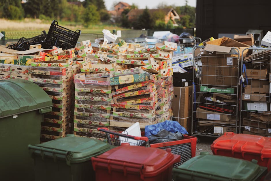 A row of large, rectangular wheelie bins lined up along the edge of a pavement beside a brick wall. The bins are made of durable plastic with textured lids and come in various colours, including green